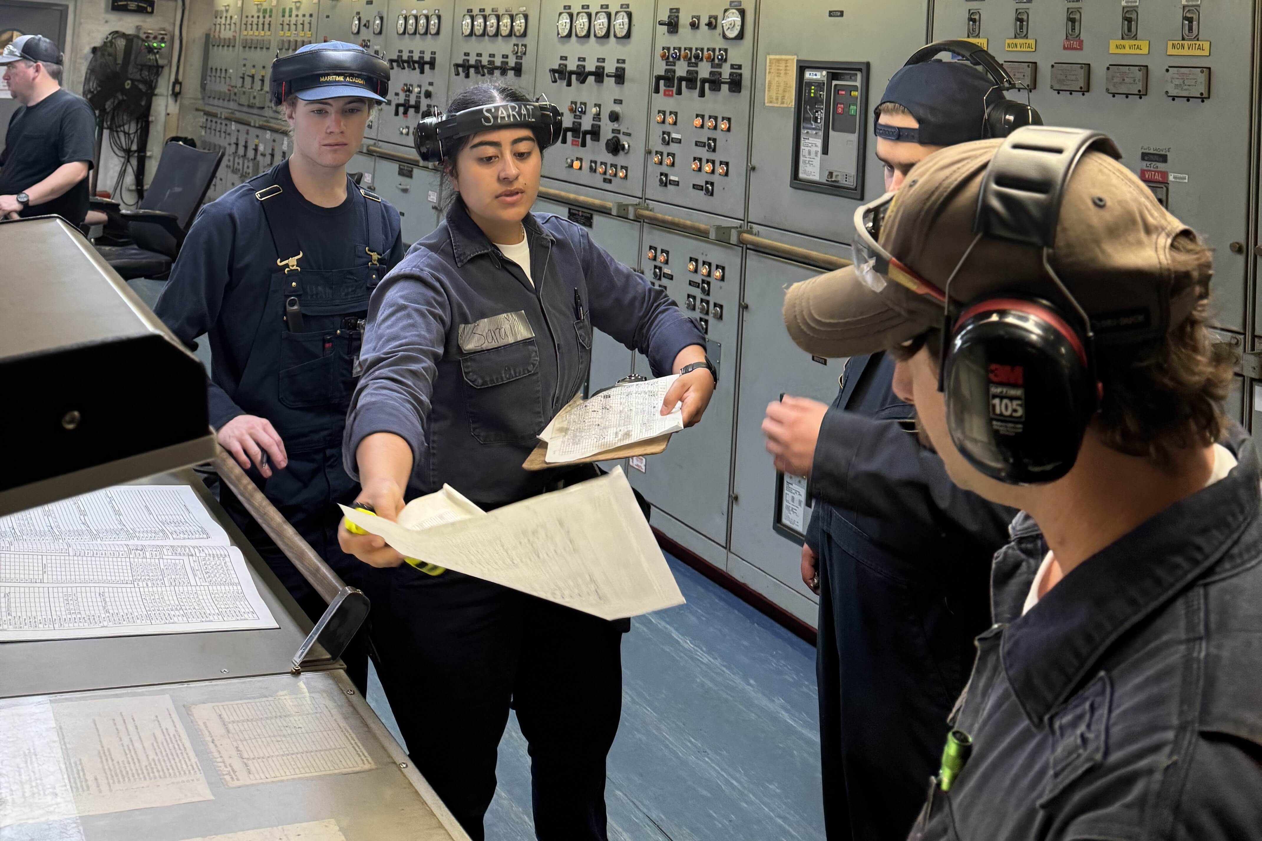 cadets wearing coveralls looking at papers in an engine room
